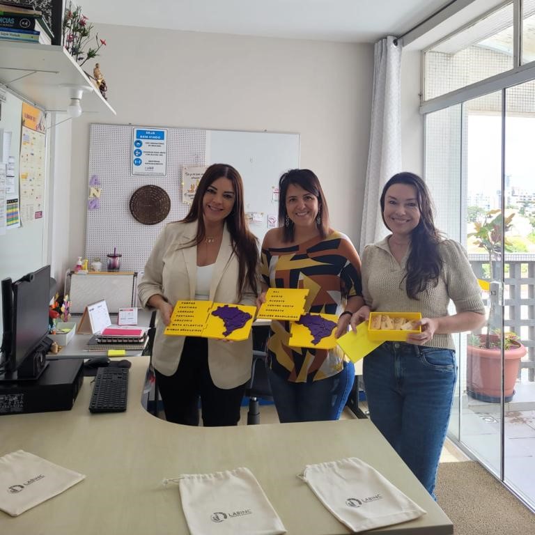 [Descrição]: Foto de uma sala pequena, com uma porta de vidro no lado direito, mesa com computador, prateleira com livros e quadros brancos com anotações em duas paredes. Sobre a mesa, em primeiro plano, há três ecobags do LABINC. No centro da foto, três mulheres seguram mapas táteis e um dominó matemático. As três estão sorridentes. [Fim da descrição]